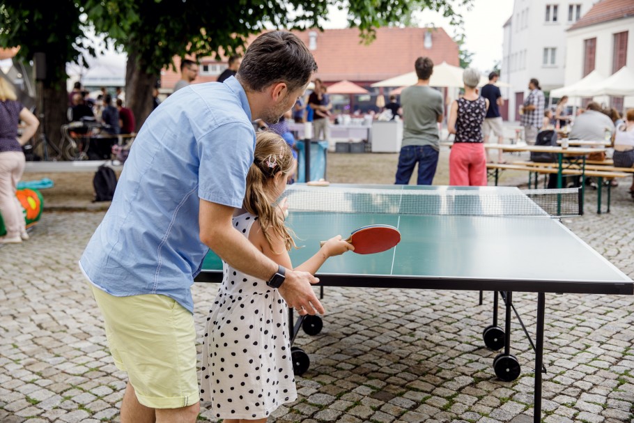 Auf unserem Sommerfest spielen ein 30 jähriger Mann und ein junges 10 jähriges Mädchen zusammen Tischtennis. Sie stehen zusammen vor der Platte, mit einem Schläger in der Hand.