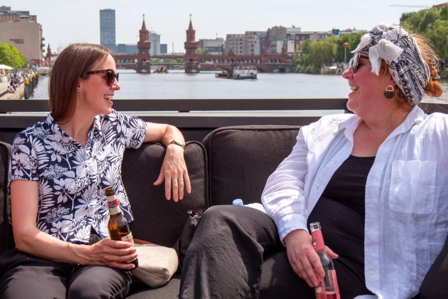 Zwei etwa 30 jährige Mitarbeiterinnen sitzen in Berlin in einem Boot und lachen. Die Sonne scheint.