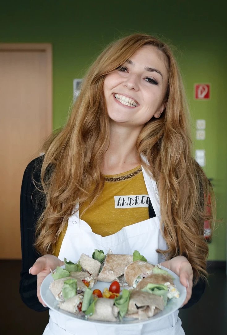 Eine etwa 20 jährige Mitarbeiterin aus Cluj hält einen Teller mit Wraps in die Kamera und lacht.