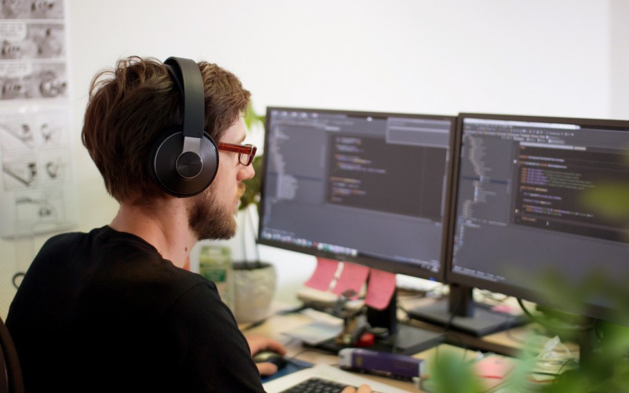 Unser 30 jähriger Mitarbeiter Sascha sitzt vor seinen zwei Monitoren an seinem Arbeitsplatz. Er hat Kopfhörer auf und widmet sich dem Programmieren.