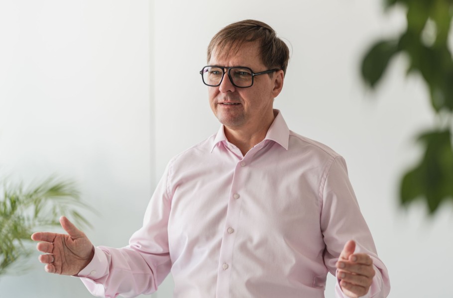 Stefan Bauer, Geschäftsführer der queo München AG, erzählt über den Zusammenschluss. Er trägt ein rosafarbenes langärmliges Hemd und eine Brille.