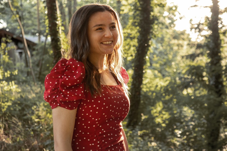 Eine junge 25-jährige Mitarbeiterin steht lächelnd unter hellem Sonnenlicht. Mitten im Wald. Sie trägt ein rotes gepunktetes Kleid.