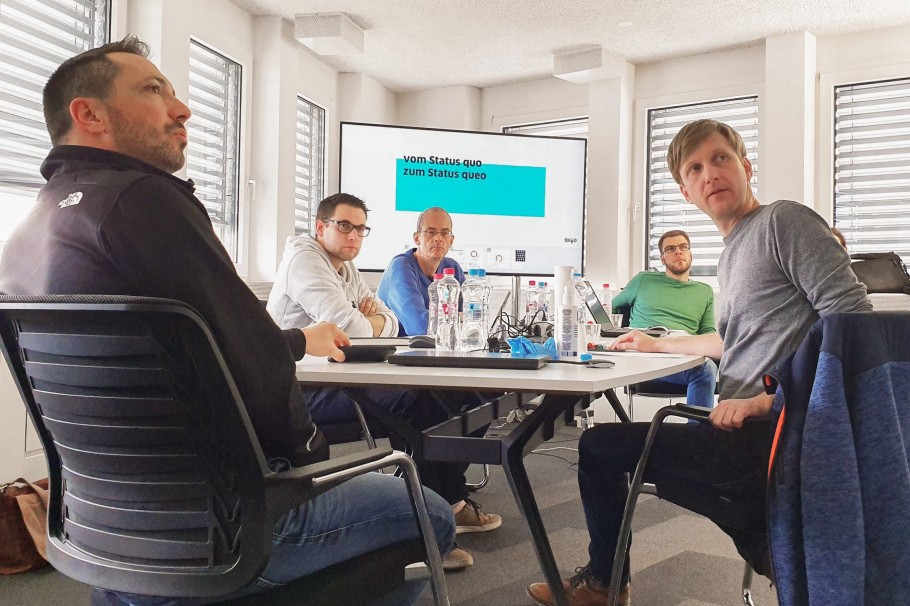 Fünf unserer Mitarbeiter sitzen beisammen an einem Konferenztisch. Hinter ihnen befindet sich ein großer Monitor. Sie schauen alle nachdenklich. Text im Bild: Vom Status quo zum Status qeuo.