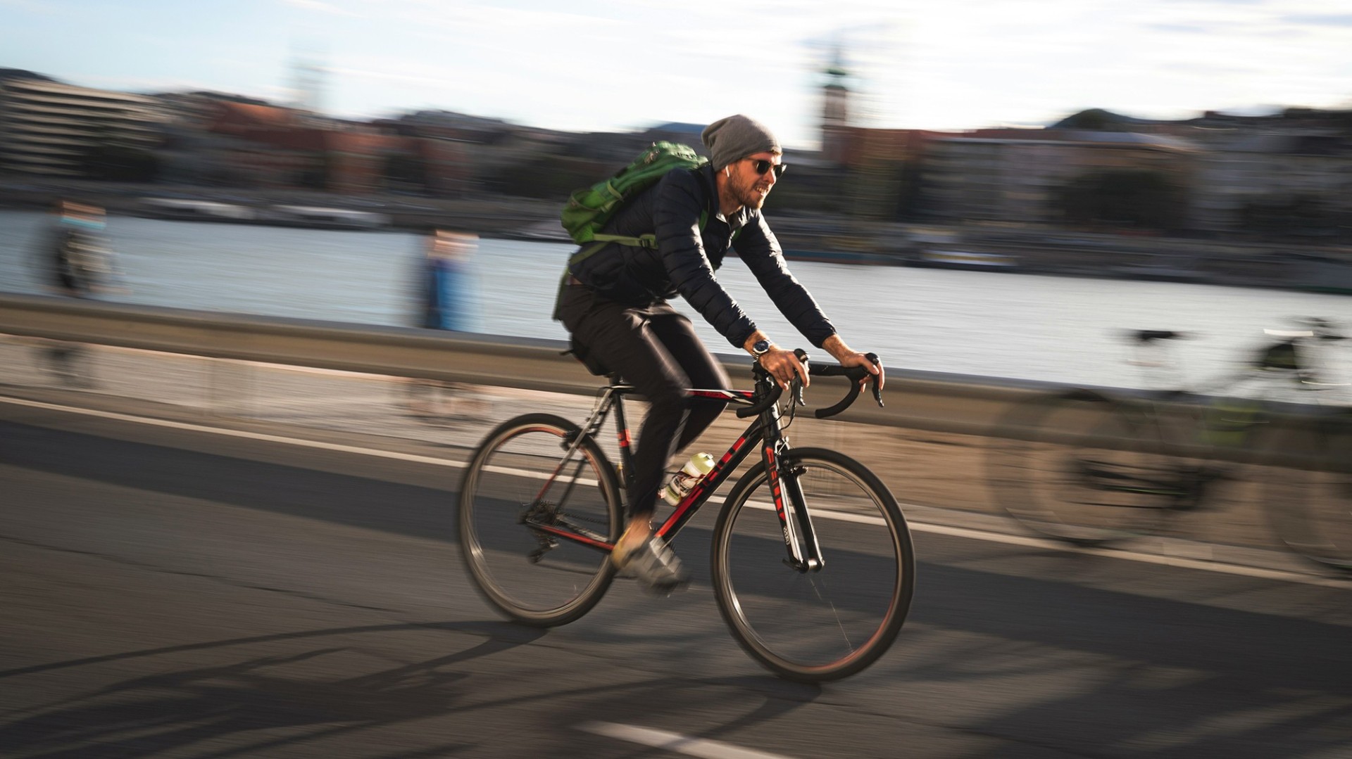 Ein 30 jähriger Mann mit Mütze und Sonnenbrille fährt mit seinem Fahrrad am Flussufer entlang. Er steht im Fokus, während seine Umgebung und andere Menschen unscharf an ihm vorbeiziehen.