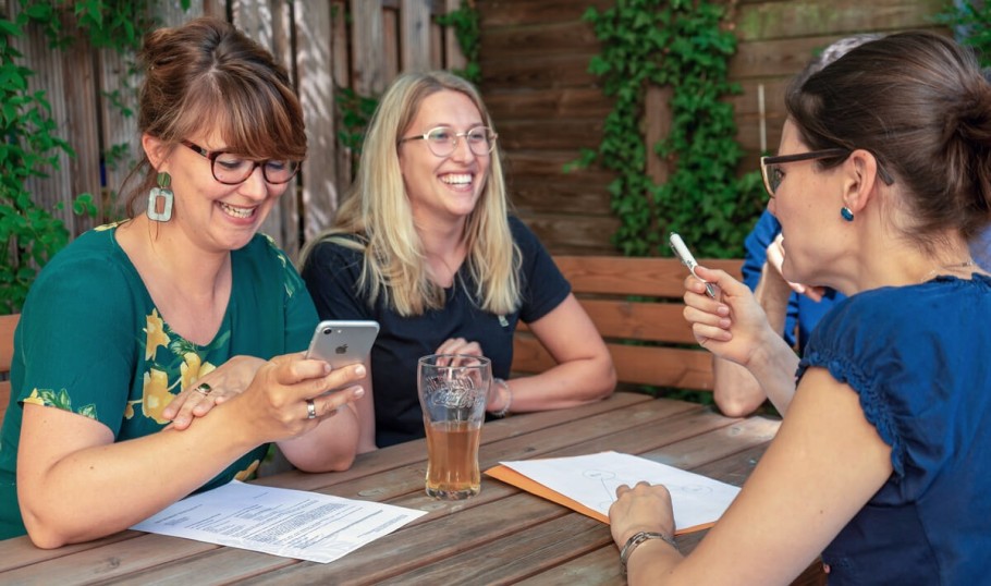 Drei 30-jährige Mitarbeiterinnen unterhalten sich lächelnd. Sie sitzen beisammen an einem großen Holztisch, auf denen Unterlagen liegen. Die links sitzende Frauen zeigt etwas auf ihrem Handy, während die Frau rechts vom Bild zustimmend mit ihrem Stift gestikuliert.