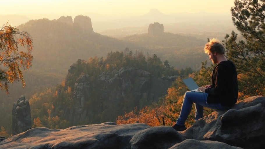 Ein etwa 30 jähriger Mann sitzt mit einem Laptop in der Sächsischen Schweiz auf einer Aussicht. Im Hintergrund sind die Schrammsteine und der Falkenstein zu sehen. Es ist Herbst. Die Sonne geht unter und Dunst liegt über dem Tal.