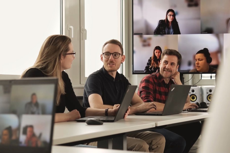 Einblick in einen Konferenzraum bei queo in Dresden. Drei Mitarbeiter sitzen nebeneinander, vor ihren Laptops. Hinter ihnen befindet sich ein großer Monitor, auf der eine Teams Besprechung eingeblendet ist.